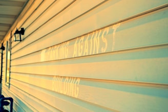 A close-up view of a building's exterior wall with horizontal siding, illuminated by warm sunlight. The faded text, partially visible, appears on the wall. Two black lantern-style outdoor lights are mounted on the wall.