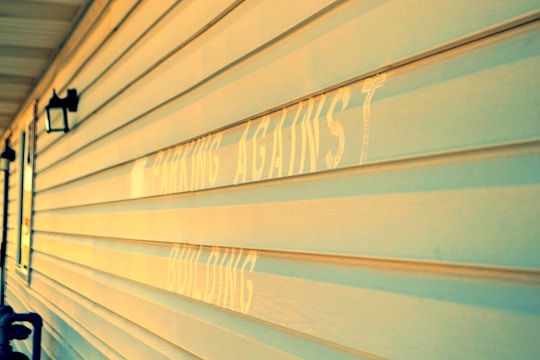 A close-up view of a building's exterior wall with horizontal siding, illuminated by warm sunlight. The faded text, partially visible, appears on the wall. Two black lantern-style outdoor lights are mounted on the wall.