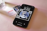A smartphone displaying a wallpaper with the image of Earth surrounded by QR code elements. It shows the time as 3:20 with the date Sunday, November 12. Next to the phone is a business card with the logo 'UNIQ R' and contact information. The setting is on a soft pink surface with a glass object partially visible.