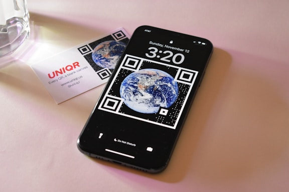 A smartphone displaying a wallpaper with the image of Earth surrounded by QR code elements. It shows the time as 3:20 with the date Sunday, November 12. Next to the phone is a business card with the logo 'UNIQ R' and contact information. The setting is on a soft pink surface with a glass object partially visible.