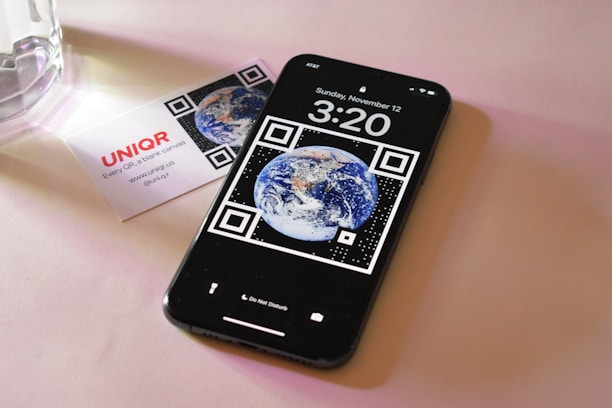 A smartphone displaying a wallpaper with the image of Earth surrounded by QR code elements. It shows the time as 3:20 with the date Sunday, November 12. Next to the phone is a business card with the logo 'UNIQ R' and contact information. The setting is on a soft pink surface with a glass object partially visible.