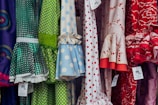 A colorful display of dresses with various polka dot and floral patterns. The fabrics feature a mix of vibrant colors including green, blue, red, and white. Ruffled sleeves and lace accents add texture, while price tags are visible hanging from some of the garments.