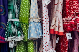 A colorful display of dresses with various polka dot and floral patterns. The fabrics feature a mix of vibrant colors including green, blue, red, and white. Ruffled sleeves and lace accents add texture, while price tags are visible hanging from some of the garments.