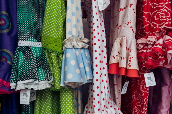 A colorful display of dresses with various polka dot and floral patterns. The fabrics feature a mix of vibrant colors including green, blue, red, and white. Ruffled sleeves and lace accents add texture, while price tags are visible hanging from some of the garments.
