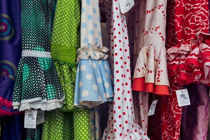 A colorful display of dresses with various polka dot and floral patterns. The fabrics feature a mix of vibrant colors including green, blue, red, and white. Ruffled sleeves and lace accents add texture, while price tags are visible hanging from some of the garments.