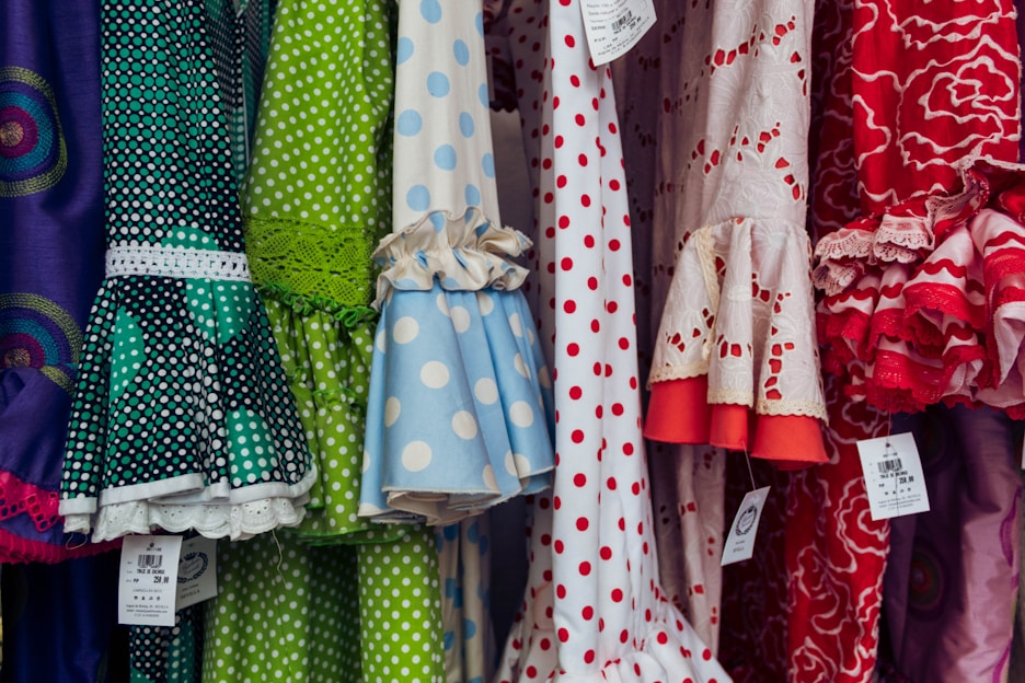 A colorful display of dresses with various polka dot and floral patterns. The fabrics feature a mix of vibrant colors including green, blue, red, and white. Ruffled sleeves and lace accents add texture, while price tags are visible hanging from some of the garments.