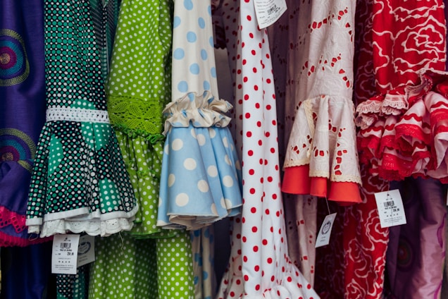 A colorful display of dresses with various polka dot and floral patterns. The fabrics feature a mix of vibrant colors including green, blue, red, and white. Ruffled sleeves and lace accents add texture, while price tags are visible hanging from some of the garments.