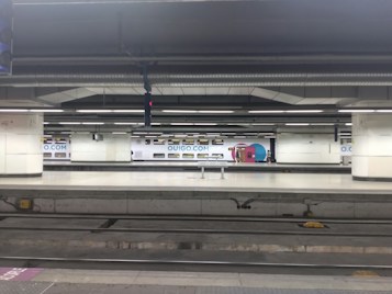 A high-speed train is stationary inside an underground train station. The train is branded with 'OUIGO.COM' in blue letters. The station features large, rectangular columns and overhead ducting. The platform is empty and the overall lighting is bright and fluorescent.