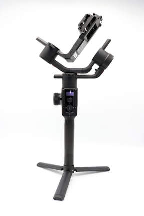 A camera gimbal stabilizer with a black, sleek design stands on a white background. It features three-axis rotation capabilities and adjustable arms for holding a camera. The base has a tripod mount for added stability.