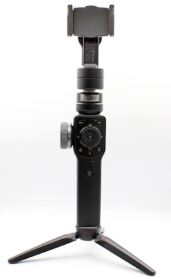 A handheld gimbal stabilizer designed for capturing steady videos and images. It features a black body with a joystick and various controls, mounted on a tripod. The holder at the top is adjustable, suitable for securing a smartphone or small camera.