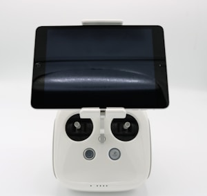 A white electronic device with a tablet screen mounted on top, featuring two control sticks and three buttons on the base. The setup is likely a remote controller for a drone or similar device, with the tablet serving as a display interface.