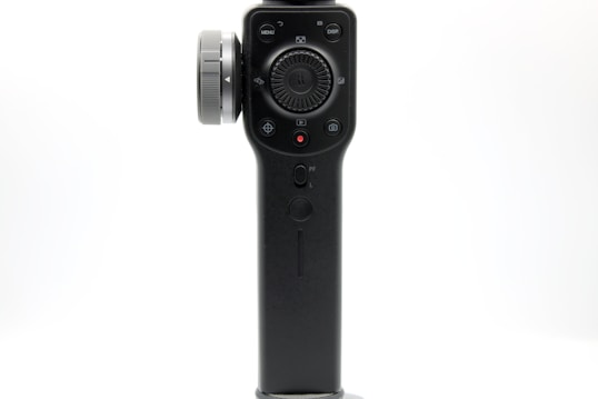 A close-up view of a black handheld electronic device with various buttons and a dial. The device features a central control panel, with buttons labeled 'MENU' and 'DISP' on the top. A large circular dial is located in the middle, surrounded by several smaller buttons and a red record button.