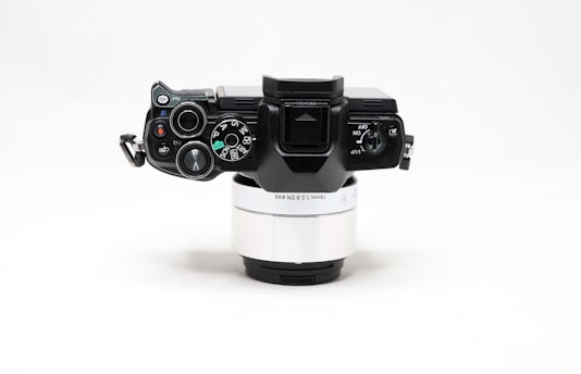 A digital camera viewed from the top, featuring various control dials and buttons, mounted on a silver lens. The body is predominantly black with detailed markings and labels on the controls, which include options like auto and manual modes.