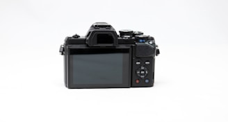 A digital camera viewed from the back showing the LCD display and various control buttons. The camera is black and positioned against a plain light background. The control buttons are arranged neatly beside the display screen, with labels such as menu, info, and other settings.