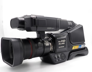 A professional video camera with a large lens, capable of 50x zoom. It features labels indicating Full HD capability, 28mm wide lens, and dual memory slots. The design includes a microphone and various control buttons, suggesting advanced recording functionalities.