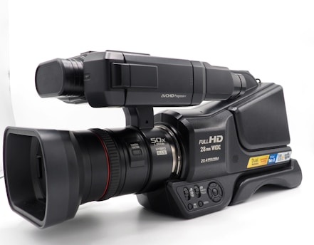 A professional video camera with a large lens, capable of 50x zoom. It features labels indicating Full HD capability, 28mm wide lens, and dual memory slots. The design includes a microphone and various control buttons, suggesting advanced recording functionalities.