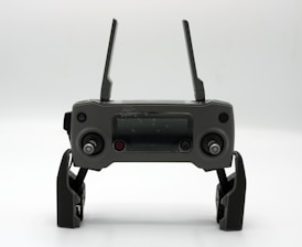 A gray drone controller with two antennas, a display screen in the center, and two control sticks on each side. The controller is positioned upright on a white surface.
