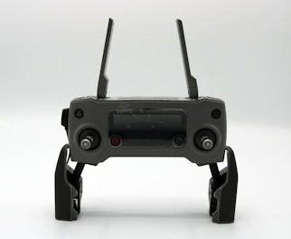 A gray drone controller with two antennas, a display screen in the center, and two control sticks on each side. The controller is positioned upright on a white surface.
