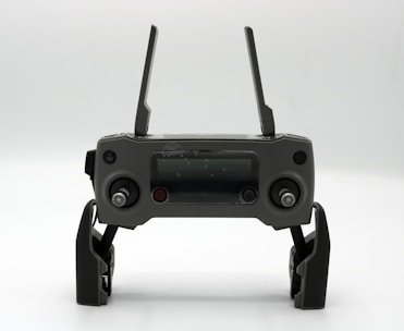 A gray drone controller with two antennas, a display screen in the center, and two control sticks on each side. The controller is positioned upright on a white surface.
