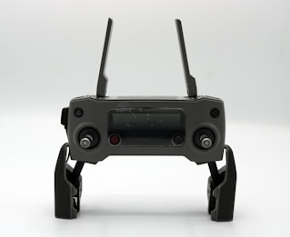 A gray drone controller with two antennas, a display screen in the center, and two control sticks on each side. The controller is positioned upright on a white surface.