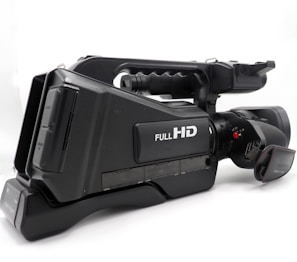 A professional video camera with a black body featuring a label that indicates Full HD capability. The camera has a large lens and a side handle for easy grip, along with several control buttons and input ports visible on the side.