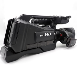 A professional video camera with a black body featuring a label that indicates Full HD capability. The camera has a large lens and a side handle for easy grip, along with several control buttons and input ports visible on the side.