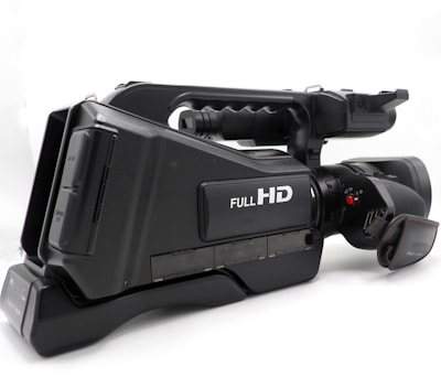 A professional video camera with a black body featuring a label that indicates Full HD capability. The camera has a large lens and a side handle for easy grip, along with several control buttons and input ports visible on the side.