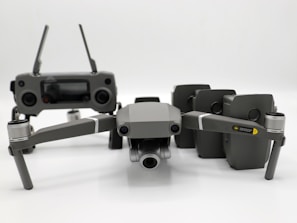 A grey drone with four rotors and a detachable camera is placed in the foreground, alongside its remote controller that has two joysticks and an LCD screen. Behind the drone, there are multiple battery packs neatly arranged.