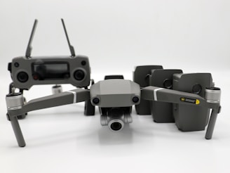 A grey drone with four rotors and a detachable camera is placed in the foreground, alongside its remote controller that has two joysticks and an LCD screen. Behind the drone, there are multiple battery packs neatly arranged.