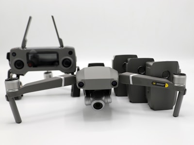 A grey drone with four rotors and a detachable camera is placed in the foreground, alongside its remote controller that has two joysticks and an LCD screen. Behind the drone, there are multiple battery packs neatly arranged.