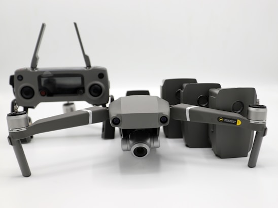 A grey drone with four rotors and a detachable camera is placed in the foreground, alongside its remote controller that has two joysticks and an LCD screen. Behind the drone, there are multiple battery packs neatly arranged.