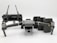 A grey drone with four rotors and a detachable camera is placed in the foreground, alongside its remote controller that has two joysticks and an LCD screen. Behind the drone, there are multiple battery packs neatly arranged.