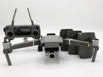 A grey drone with four rotors and a detachable camera is placed in the foreground, alongside its remote controller that has two joysticks and an LCD screen. Behind the drone, there are multiple battery packs neatly arranged.