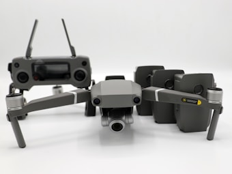 A grey drone with four rotors and a detachable camera is placed in the foreground, alongside its remote controller that has two joysticks and an LCD screen. Behind the drone, there are multiple battery packs neatly arranged.