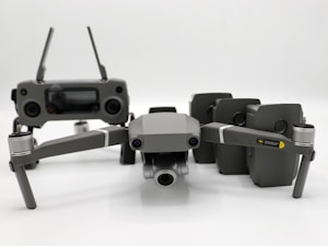 A grey drone with four rotors and a detachable camera is placed in the foreground, alongside its remote controller that has two joysticks and an LCD screen. Behind the drone, there are multiple battery packs neatly arranged.