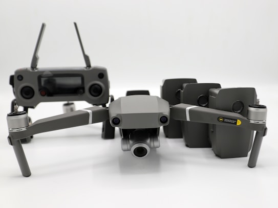 A grey drone with four rotors and a detachable camera is placed in the foreground, alongside its remote controller that has two joysticks and an LCD screen. Behind the drone, there are multiple battery packs neatly arranged.