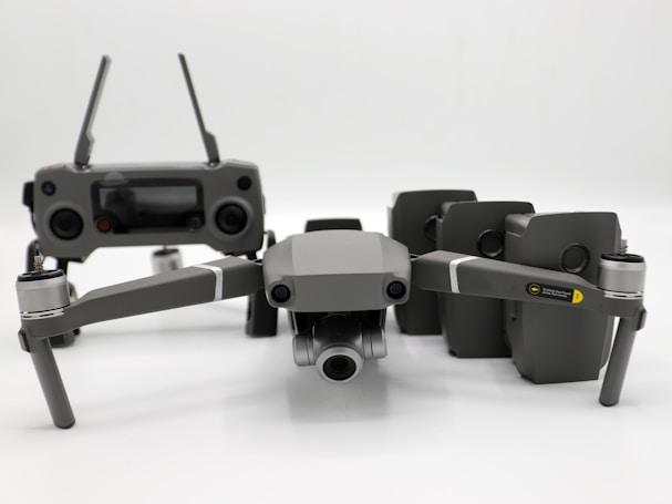A grey drone with four rotors and a detachable camera is placed in the foreground, alongside its remote controller that has two joysticks and an LCD screen. Behind the drone, there are multiple battery packs neatly arranged.