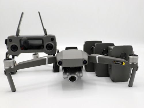 A grey drone with four rotors and a detachable camera is placed in the foreground, alongside its remote controller that has two joysticks and an LCD screen. Behind the drone, there are multiple battery packs neatly arranged.