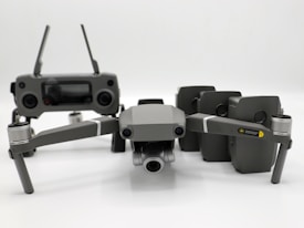 A grey drone with four rotors and a detachable camera is placed in the foreground, alongside its remote controller that has two joysticks and an LCD screen. Behind the drone, there are multiple battery packs neatly arranged.