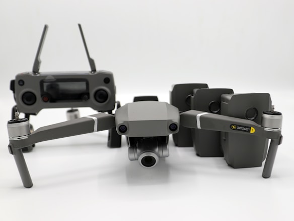 A grey drone with four rotors and a detachable camera is placed in the foreground, alongside its remote controller that has two joysticks and an LCD screen. Behind the drone, there are multiple battery packs neatly arranged.