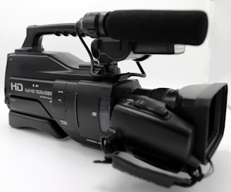 A professional video camera with a large microphone attached on top, featuring a handle for easy carrying and multiple control buttons for functionality. The camera is predominantly black and has a label indicating full HD capability with 1920x1080 resolution.