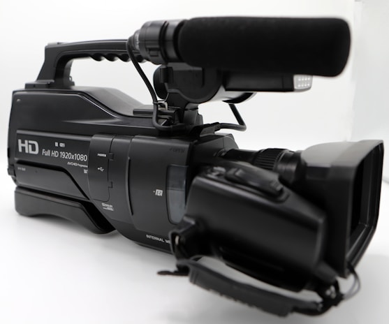 A professional video camera with a large microphone attached on top, featuring a handle for easy carrying and multiple control buttons for functionality. The camera is predominantly black and has a label indicating full HD capability with 1920x1080 resolution.