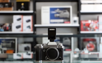 A classic-style camera with a metallic body and textured grip sits prominently on a glass surface. The camera's flash is extended, with a backdrop of a blurred store display featuring various photography and electronic equipment boxes.