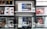 A store shelf filled with various boxed electronic items, including lenses and cameras, arranged in an orderly fashion. In the foreground, there is an empty frame-like object, possibly a camera accessory, placed on a reflective glass surface.