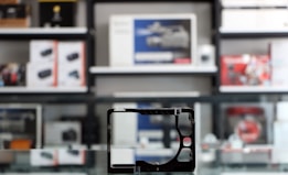 A store shelf filled with various boxed electronic items, including lenses and cameras, arranged in an orderly fashion. In the foreground, there is an empty frame-like object, possibly a camera accessory, placed on a reflective glass surface.