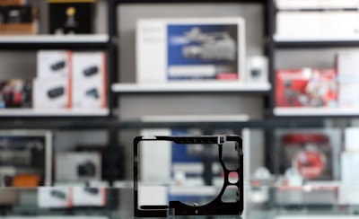 A store shelf filled with various boxed electronic items, including lenses and cameras, arranged in an orderly fashion. In the foreground, there is an empty frame-like object, possibly a camera accessory, placed on a reflective glass surface.