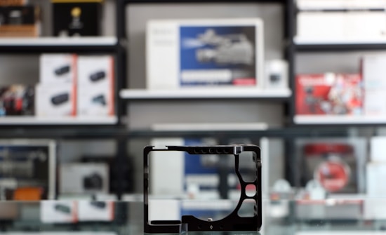 A store shelf filled with various boxed electronic items, including lenses and cameras, arranged in an orderly fashion. In the foreground, there is an empty frame-like object, possibly a camera accessory, placed on a reflective glass surface.