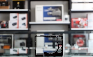 A store shelf filled with various boxed electronic items, including lenses and cameras, arranged in an orderly fashion. In the foreground, there is an empty frame-like object, possibly a camera accessory, placed on a reflective glass surface.