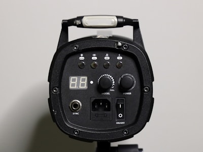A close-up view of a black electronic device, possibly a studio light or strobe, featuring various control buttons and a digital display. The device has knobs labeled 'Model' and 'Power', along with buttons for 'Beep' and 'Test'. An on/off switch and a sync port are also visible, as well as a carry handle at the top.