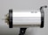 A silver studio light with black accents mounted on a support bracket. The cylindrical shape features small vents and a clear bulb extending from the left side.